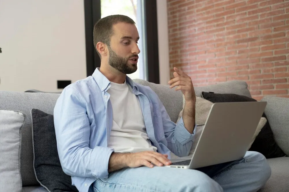 homem no sofá usando laptop enquanto fala durante uma videochamada em casa cena de trabalho remoto e teletrabalho casual com postura relaxada em uma sala de estar moderna com fundo de parede de tijolos