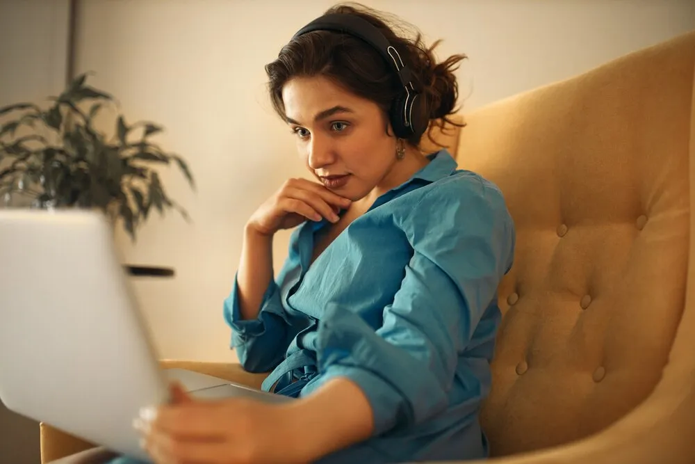bela jovem usando headset sem fio enquanto assiste a um webinar sobre marketing de mídia social, com um olhar focado. garota bonita aprendendo online sentada no sofá com um laptop, com a mão no queixo