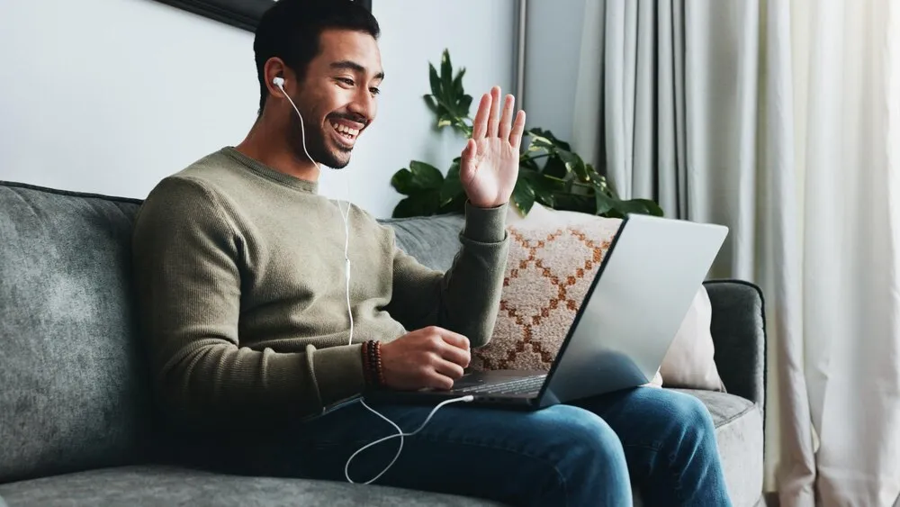 chamada de vídeo com aceno e laptop com homem no sofá para networking de freelancer e comunicação de saudação de boas-vindas e trabalhador remoto com pessoa na sala de estar de casa para pequenos negócios e streaming