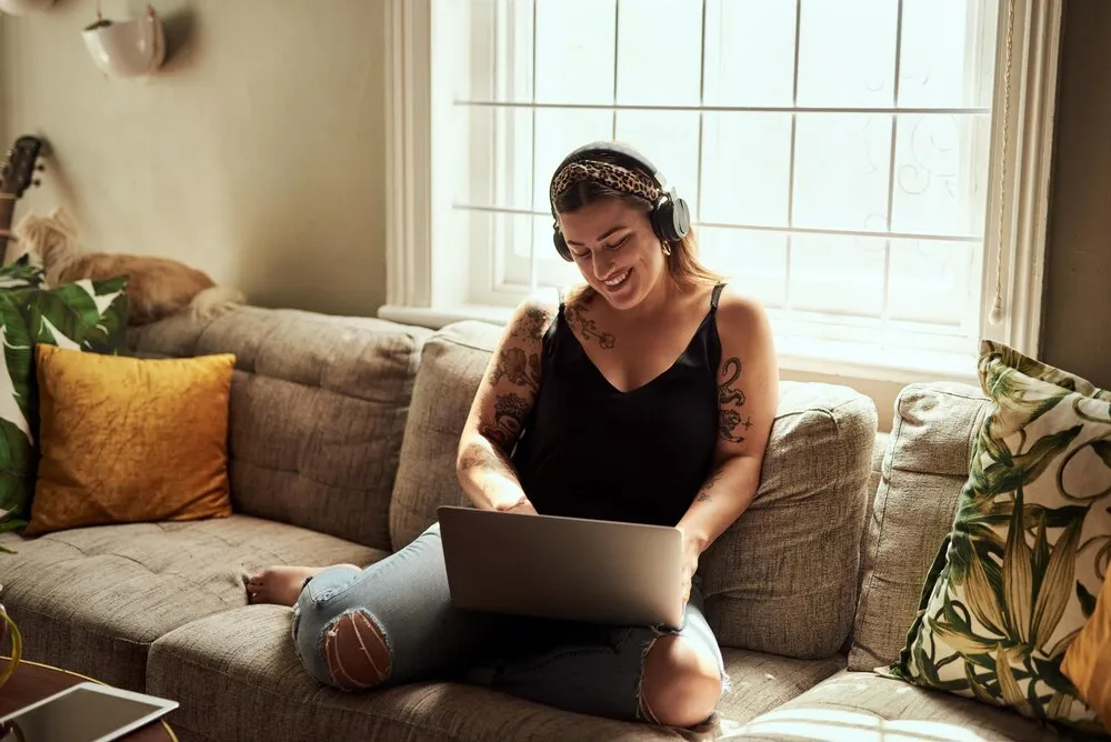 fones de ouvido de computador e mulher no sofá e música feliz trabalho em casa para saúde mental em apartamento pessoa jovem relaxando no sofá e ouvindo tecnologia de áudio eletrônicos e trabalhando no laptop