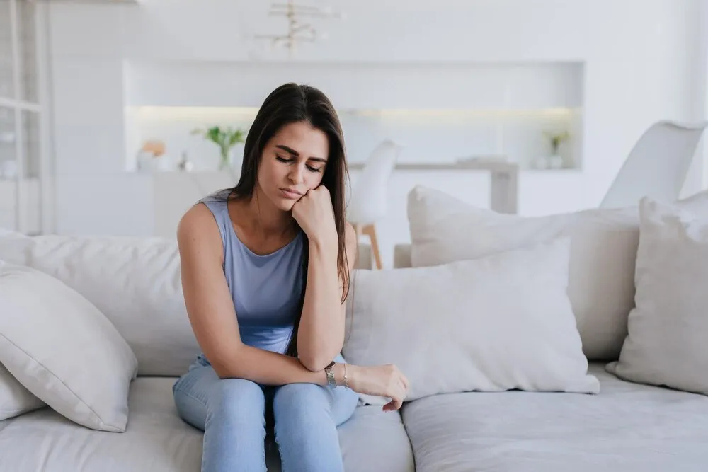 garota espanhola chateada de blusa lilás e calça jeans azul sentada no sofá em casa com os olhos voltados para baixo