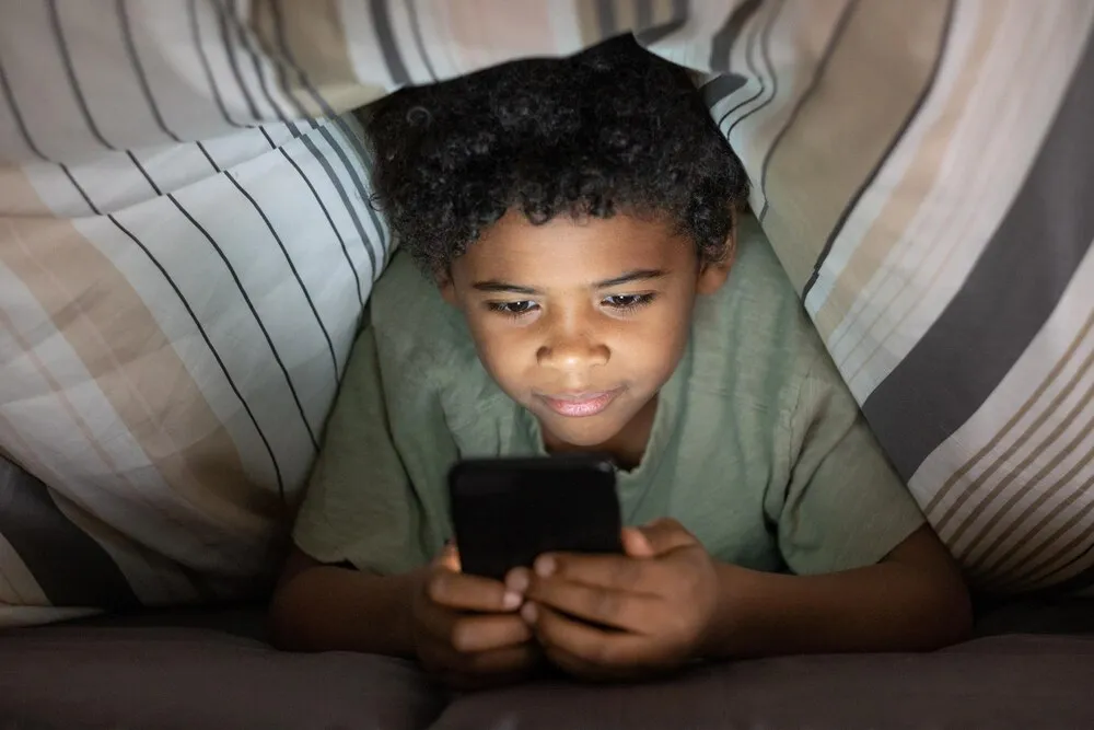 menino negro fofo e contente com cabelo cacheado deitado na cama e usando smartphone sob o edredom à noite